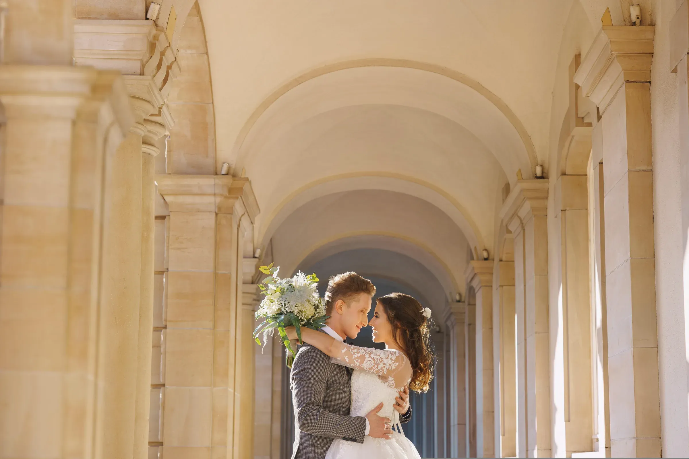 A bride and groom holding each other under a long stone colonnade, her bouquet over his shoulder.