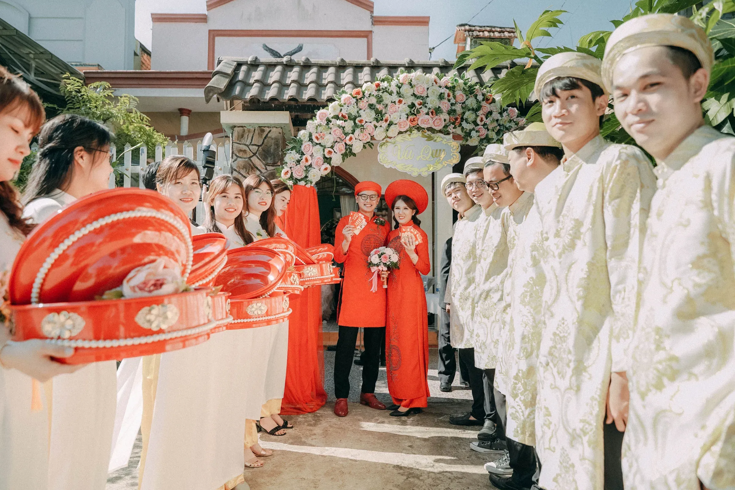 A bride and groom in red under a flower arch, with two lines of guests in white holding red gift trays.