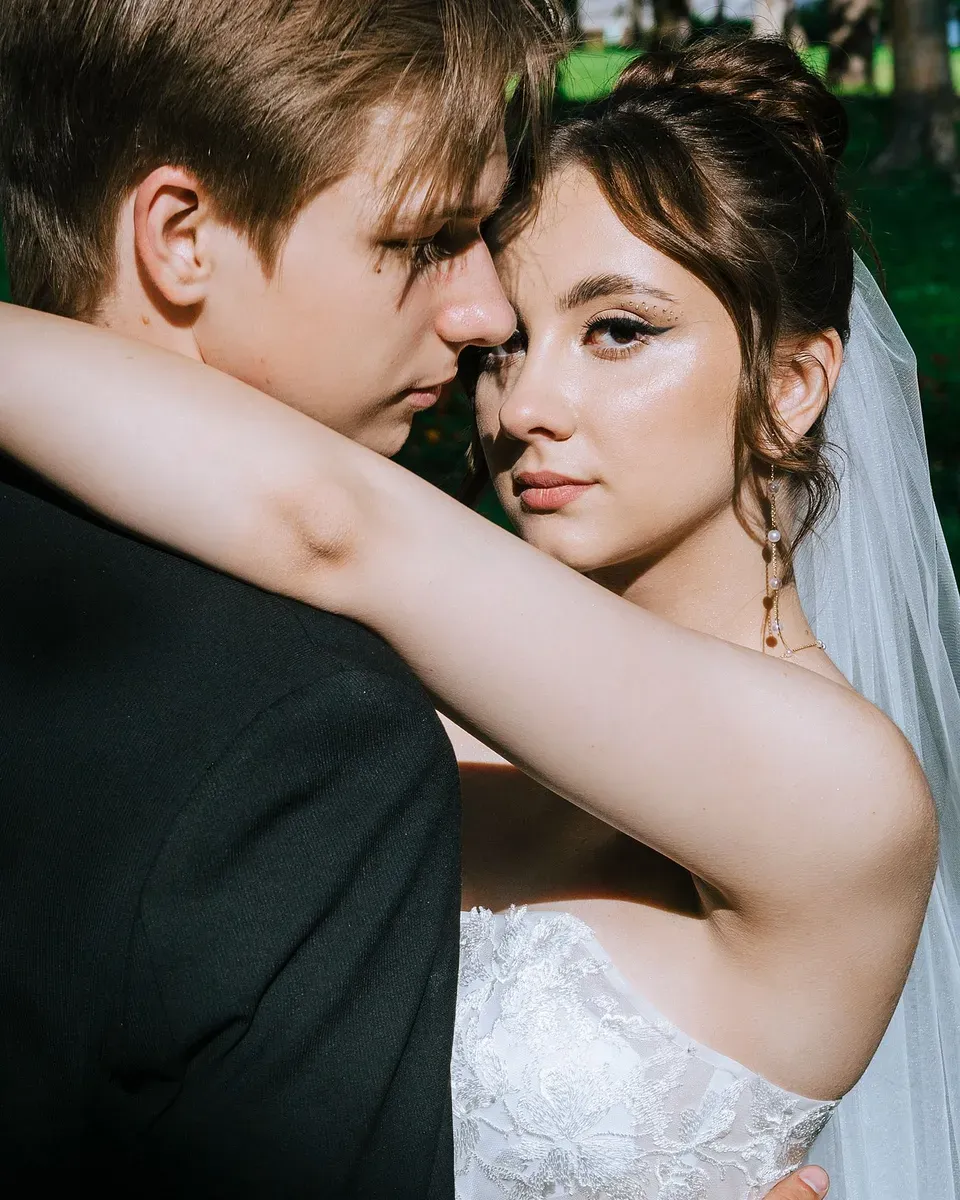 A bride and groom close together, her arm around his neck, looking at the camera.