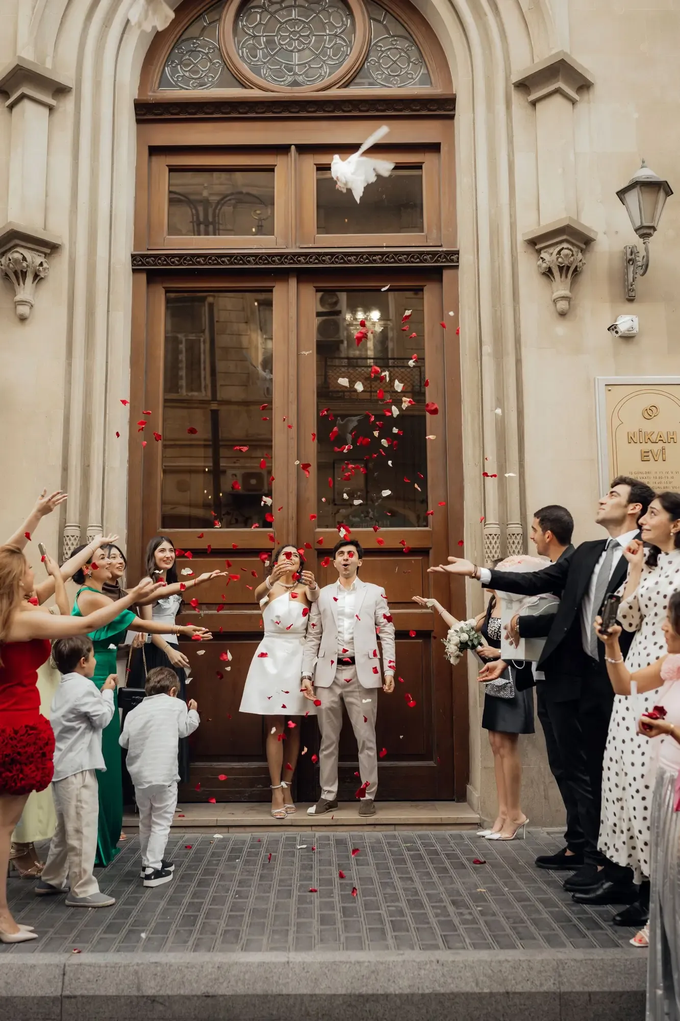 Bride and groom celebrate their wedding outside a historic building with guests throwing petals.