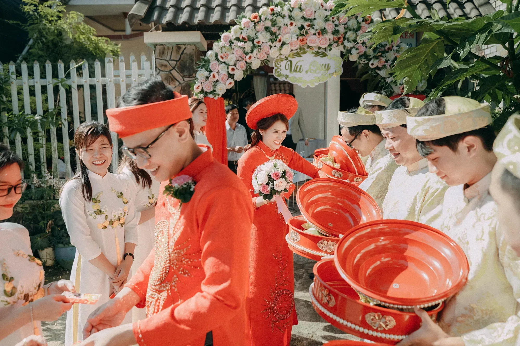 Guests in white and gold pass red lacquered trays to one another at the gate, the couple in red behind them.