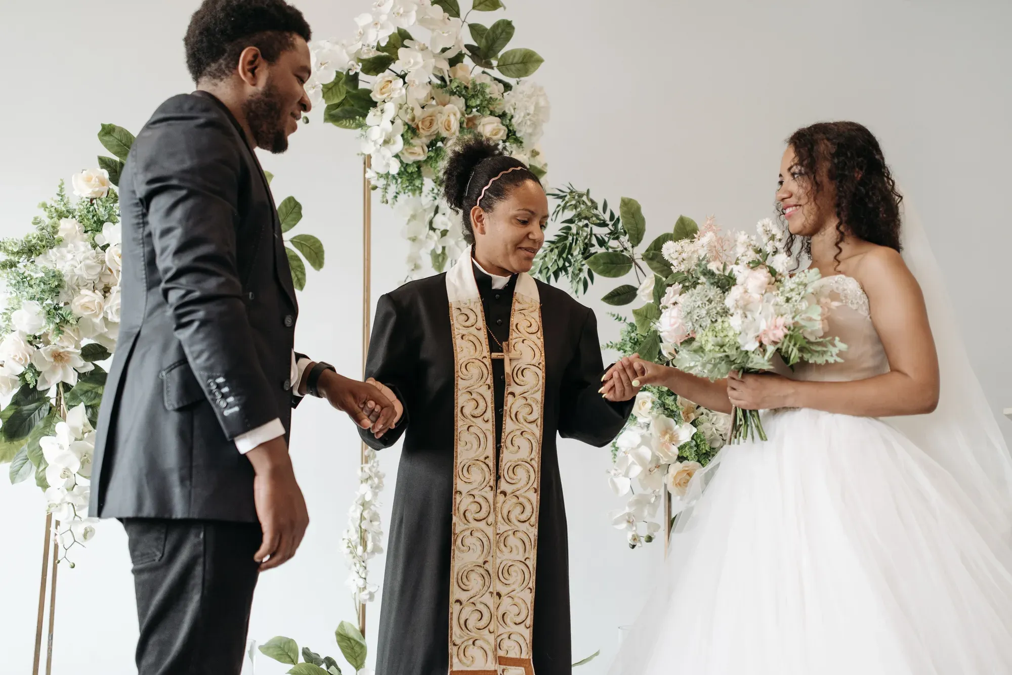 The pastor holding the bride's and groom's hands together at the front of a white room.