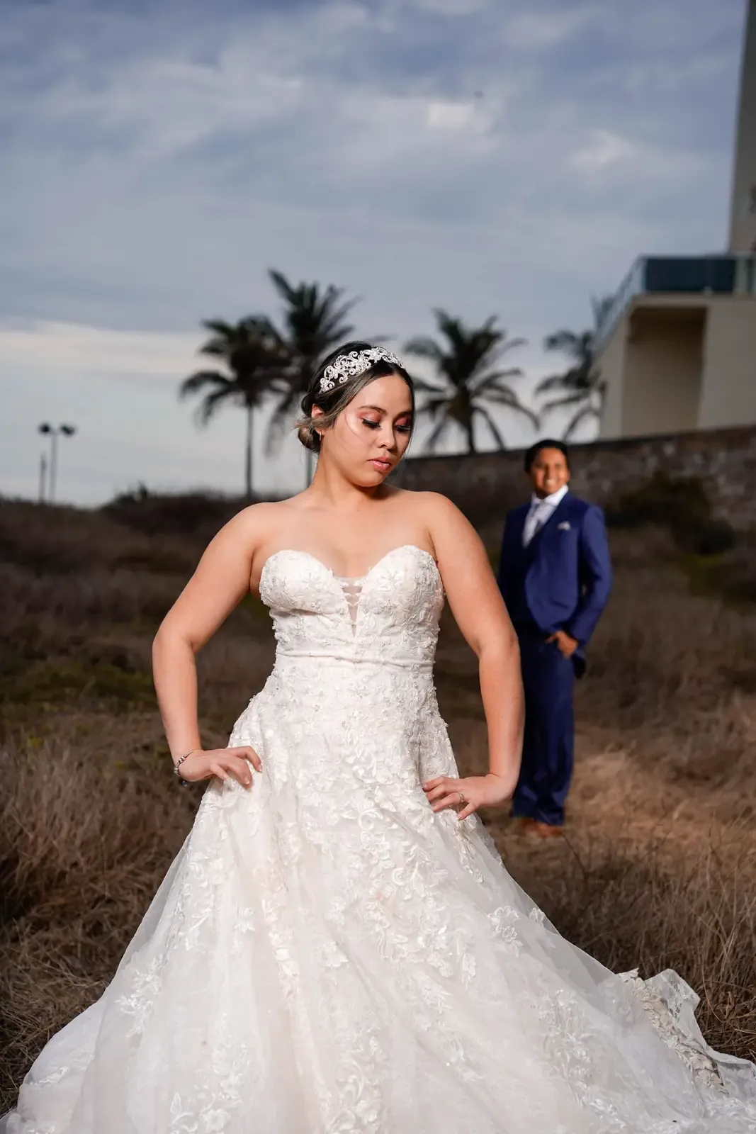 Bride in a lace gown at sunset, the groom behind her.