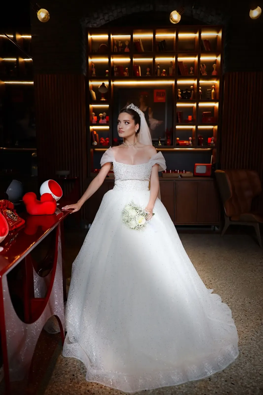 A beautiful bride in a white gown holds a bouquet indoors, with stylish decor.