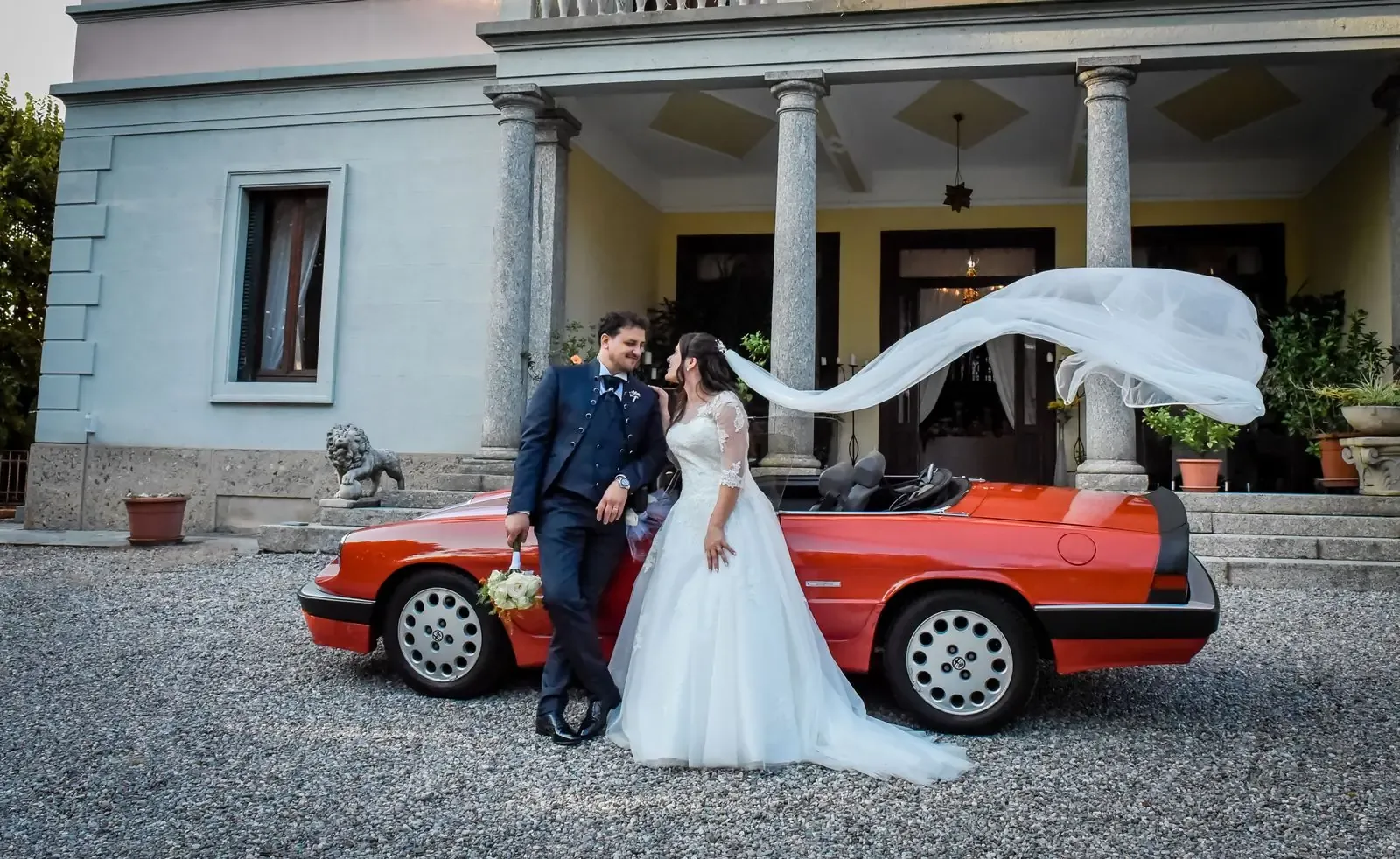 Newlyweds share a moment next to a classic red car in Como, Italy.