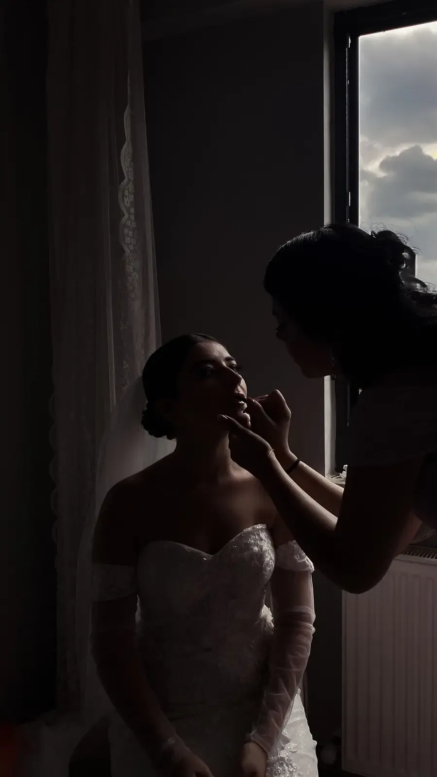 A bride getting her makeup done near a window, highlighting elegance and preparation before her wedding.