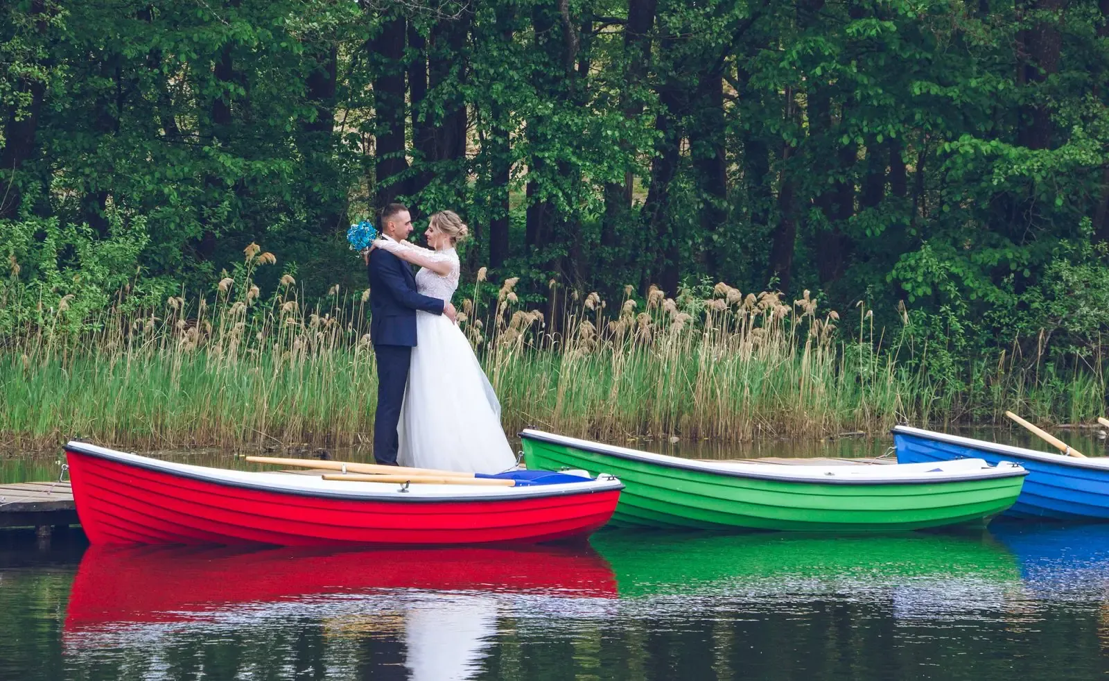 A couple embraces on colorful boats by a lush forest, celebrating their wedding day.
