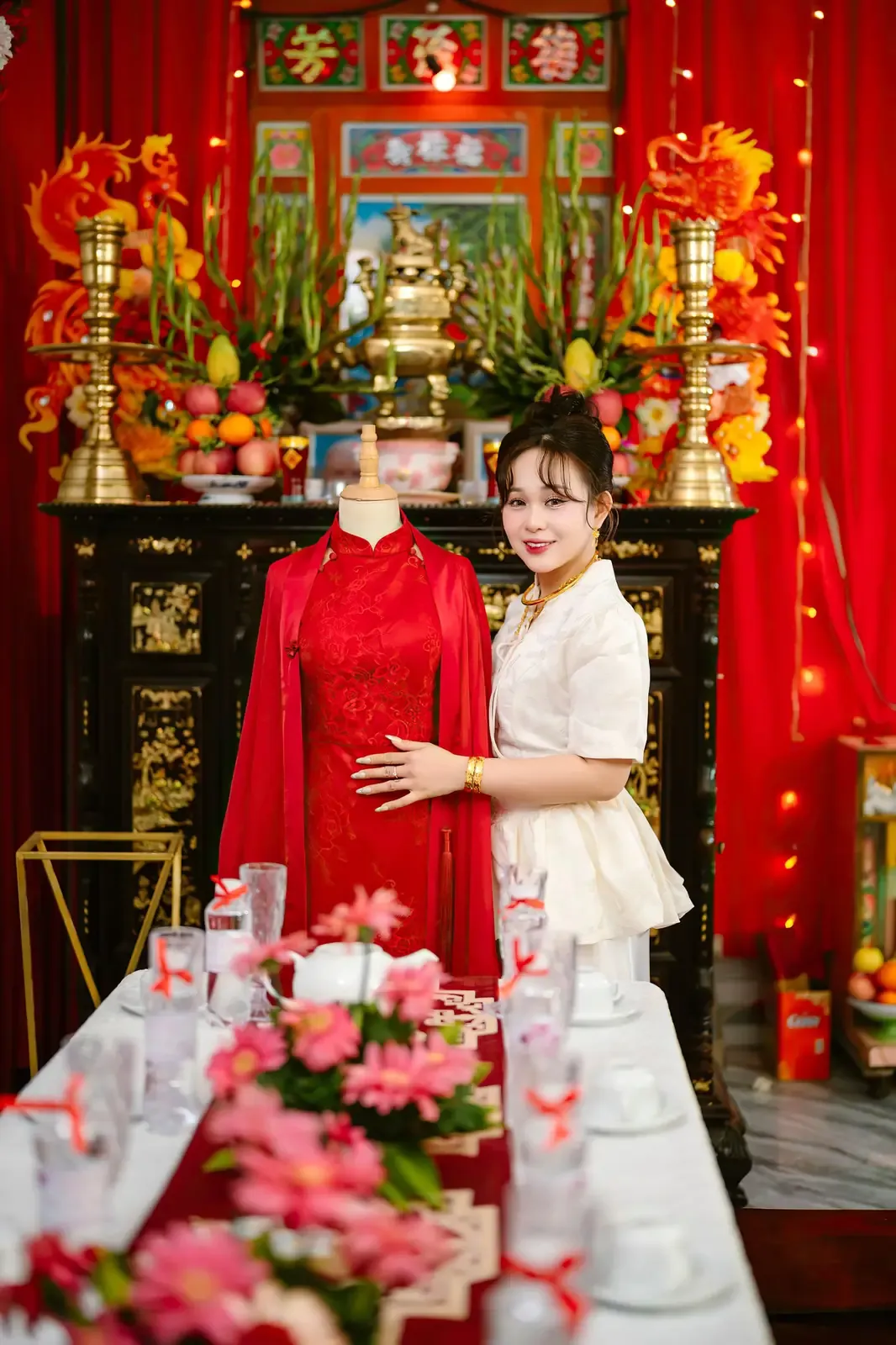 A woman stands beside a traditional Vietnamese wedding dress in a brightly decorated room.