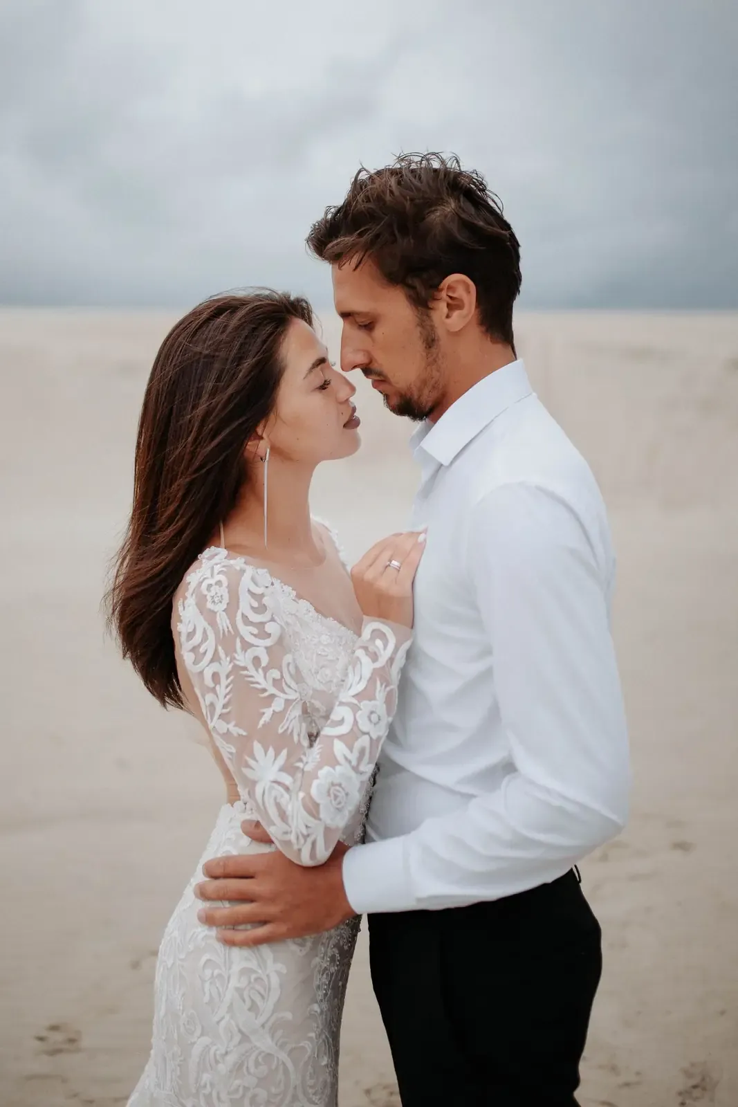 Bride and groom gently embracing on a serene beach with cloudy sky.