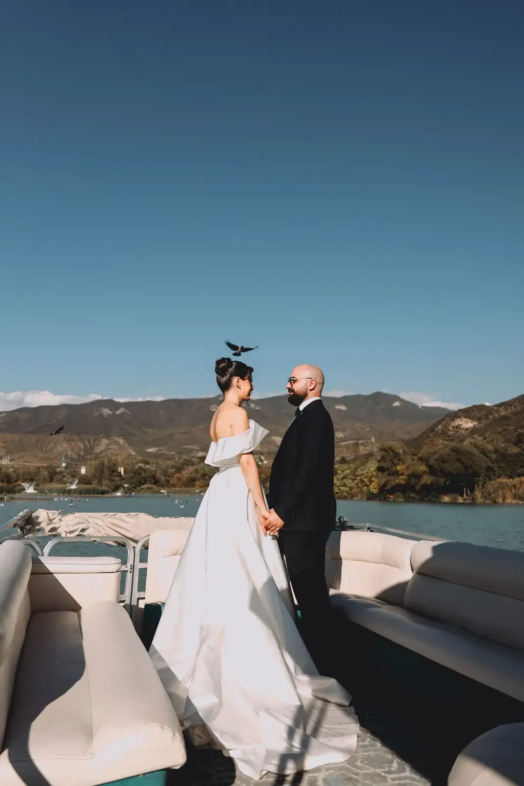 Bride and groom standing on a boat holding hands with mountains in the background.