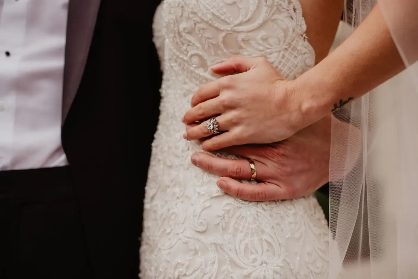 Close-up of a bride and groom embracing with focus on wedding rings and intricate dress details.