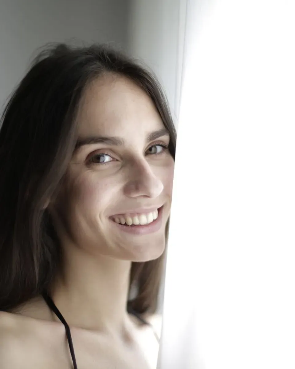 A woman smiling beside a window in soft light.
