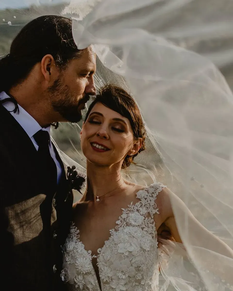 A groom kisses his bride on the forehead, her veil lifted by the wind.